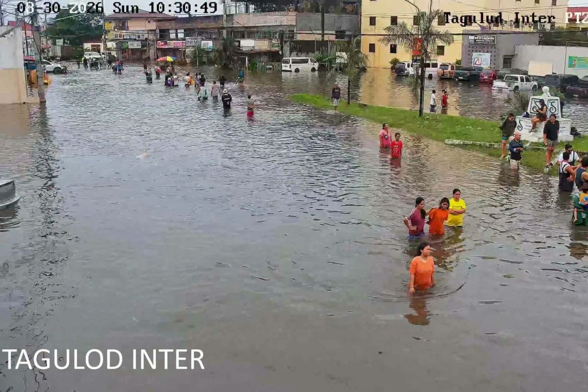 A traffic-camera view of a flooded intersection in San Fernando, dozens of residents wading through waist-deep water past half-submerged vehicles.
