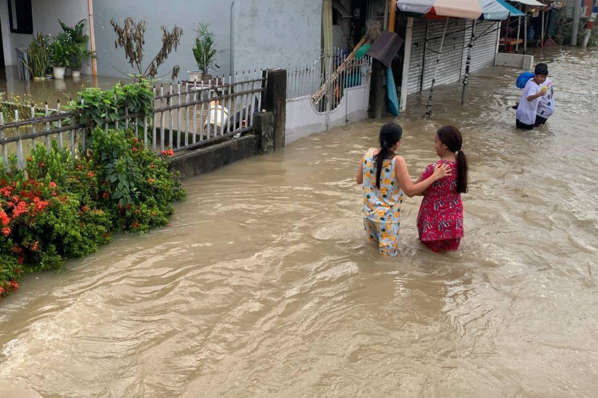 Two women wade waist-deep through muddy floodwater on a residential street in San Luis, one steadying the other by the shoulder.